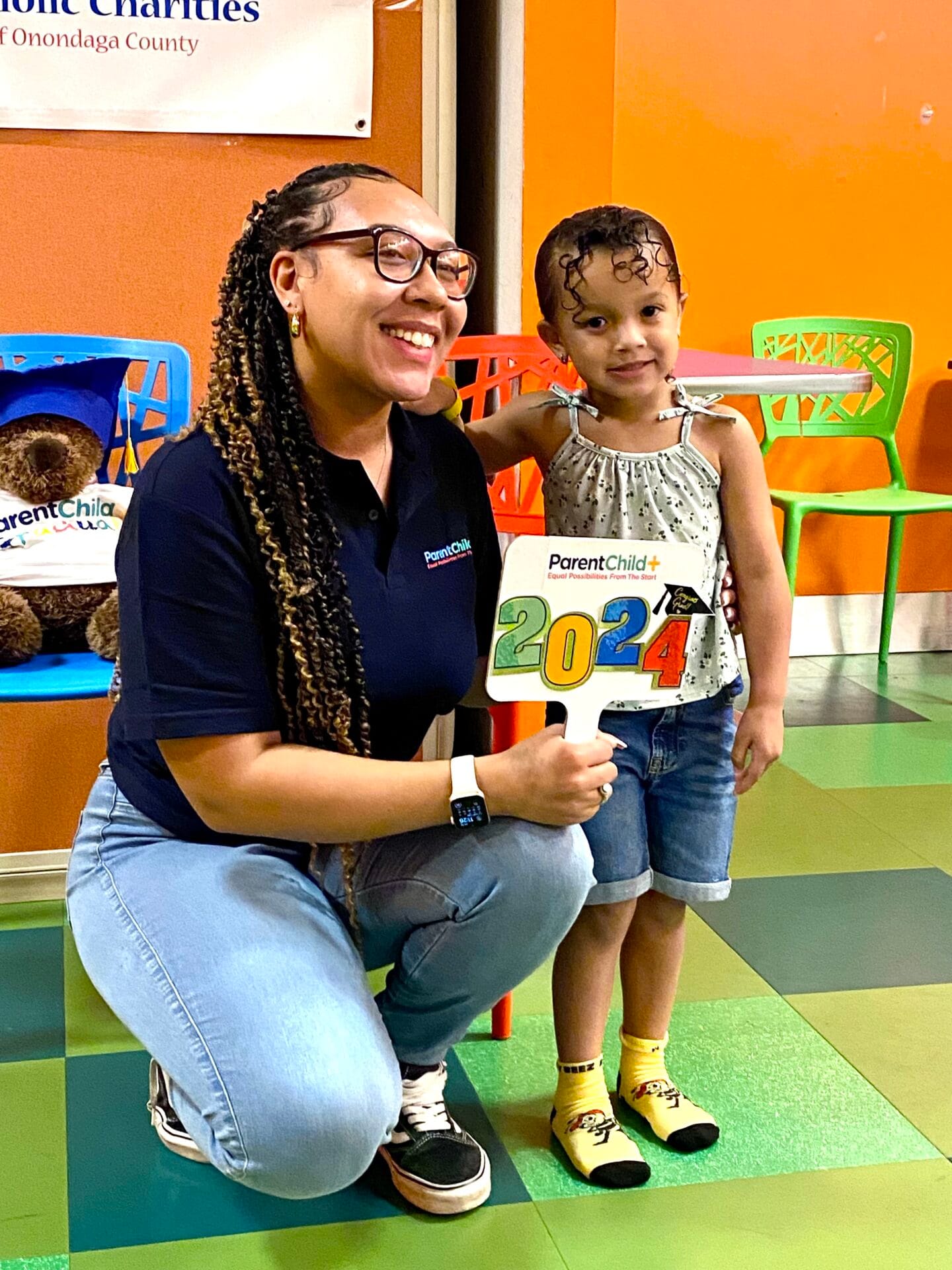 An Early Learning Specialist poses kneeling next to a child who graduated from the program in 2024. They are smiling together and attending the graduation celebration.